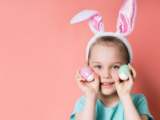 Little blonde child in headband with rabbit ears, dressed in blue t-shirt. She is smiling, showing two colorful eggs, posing against pink background. Happy Easter, childhood. Close up, copy space