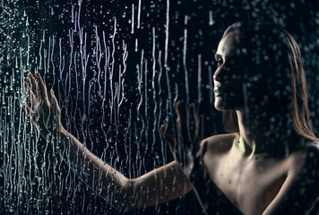 Fototapeta na wymiar A beautiful slim nude girl sensually touches with her hands a window glass, on which streams and drops of rain water flow down on a black background. Artistic design.