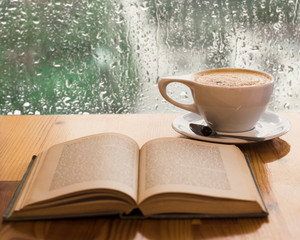 Open book and cup of coffee in front of a rainy window