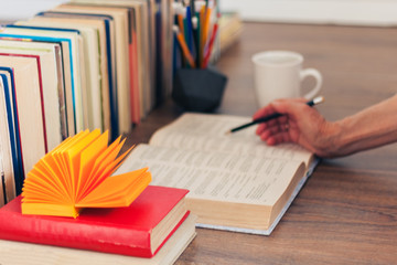 Hand of teacher, open textbook, notebook, glasses, pencils in holder, cup of tea and stack of old book on wooden table, education concept background, many books piles with copy space for text.