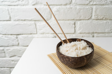 White rice in a coconut bowl on a white background. Minimalistic photo with rice and bamboo chopsticks.