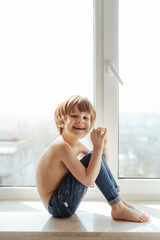 Topless kid portrait near the window. Sunny day outside. Bright white room. Boy poses, smiles and having fun alone. 
