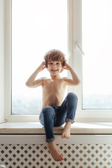 Topless kid portrait near the window. Sunny day outside. Bright white room. Boy poses, smiles and having fun alone. 
