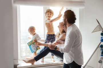 Fototapeta premium Happy smiling modern caucasian family playing and spending time in a bright white room near the window. Sunny day. Perfect parenting relationship. Parents of two cute boys.