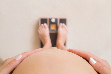 Young pregnant woman standing on weight scales. Care about body. Hands touching naked big belly....
