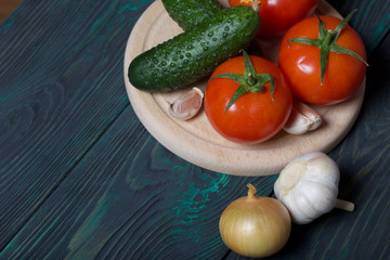 Cucumbers, tomatoes, onions and garlic on a cutting board. Tomatoes with green ponytails. Cutting board lies on the surface of brushed pine boards.