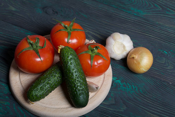 Cucumbers, tomatoes, onions and garlic on a cutting board. Tomatoes with green ponytails. Cutting board lies on the surface of brushed pine boards.