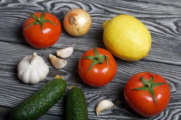 Tomatoes with green ponytails, onion, lemon and garlic head. Two green prickly cucumbers with yellow flowers. They lie on the surface of brushed pine boards.