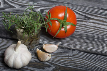 Head of garlic, a few cloves of garlic. A glass with rosemary branches. Red tomato with a green ponytail. They lie on the surface of brushed pine boards.