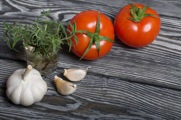 Head of garlic, a few cloves of garlic. A glass with rosemary branches. Red tomato with a green ponytail. They lie on the surface of brushed pine boards.