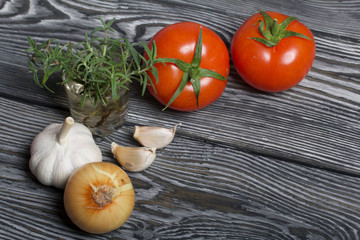 Head of garlic, a few cloves of garlic. A glass with rosemary branches. Red tomato with a green ponytail and onion. They lie on the surface of brushed pine boards.