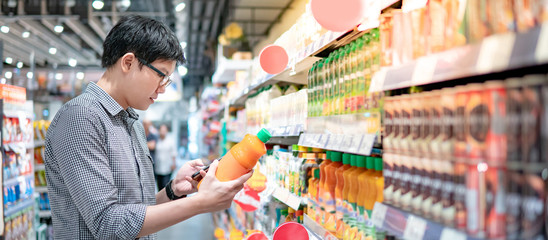 Asian man choosing orange juice in supermarket using smartphone to check shopping list. Male...