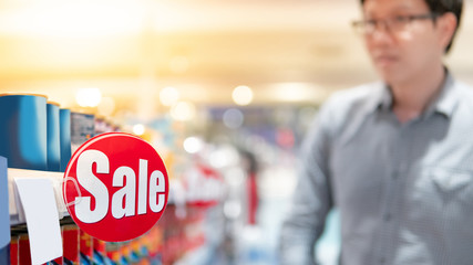 Red sale label on product shelf in supermarket with blurred male shopper choosing food package in...