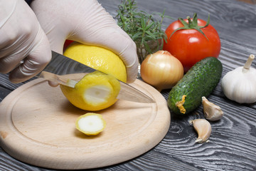 A man in rubber gloves cuts a lemon with a knife on a cutting board. Nearby are cucumber, tomato, rosemary and garlic. On the surface of brushed pine boards.