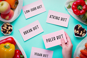 Woman choosing between popular diets for weight loss with fresh fruits, nuts and vegetables in background. Stickers with popular diet names on blue, healthy lifestyle concept, flat lay, top view - obrazy, fototapety, plakaty