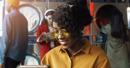 Pretty and happy African American girl in yellow glasses watching funny video on tablet device, resting and waiting for clothes to be washed. Stylish woman sipping using computer in laundry service.