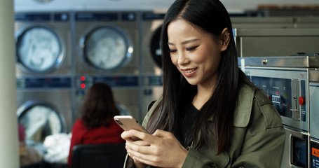 Asian pretty woman with long dark hair texting message on smartphone while standing in laundry service room. Beautiful woman typing on phone and waiting for clothes to wash in public laundromat.