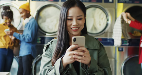 Asian young pretty and stylish girl in yellow glasses standing in laundry service room and tapping on smartphone. Woman texting message on phone while waiting for clothing to be washed.