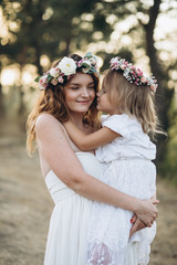 Happy elegant mother and her beautiful daughter with flower wreaths in a park at sunset