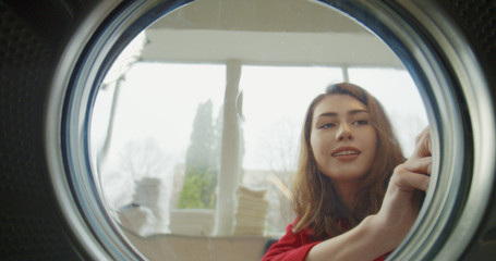 Fototapeta na wymiar View from inside washing machine on Caucasian beautiful happy woman opening up and taking out clean washed clothes. Close up of charming joyful girl in a public laundromat