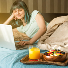 Obraz na płótnie Canvas A young woman in pajamas lies on the bed, types on a laptop. On a tray is a bowl of granola, fruits, glass with orange juice. Sunny morning for a Healthy digital breakfast and remote work from home.