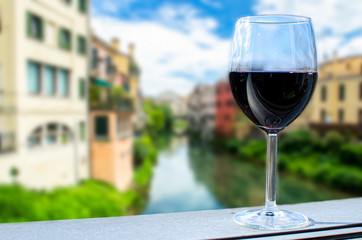 Glass of wine with canal and colorful buildings blur background, Italy. Traditional canal street with bright colorful buildings, green bushes and blue sky in Padova, Italy. Vibrant Italian cityscape.