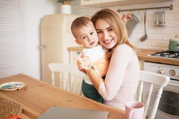 Family bonds, togetherness, love and happiness concept. Charming young Caucasian blonde mom on maternity leave embracing her cute one year old son after breakfast in stylish kitchen, smiling