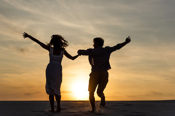 silhouettes of man and woman running on beach against sun during sunset