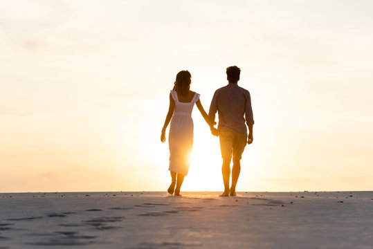 Silhouettes Of Man And Woman Holding Hands While Walking On Beach Against Sun During Sunset