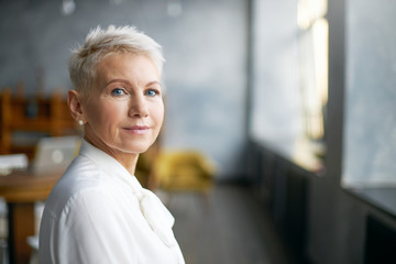 Isolated image of beautiful stylish middle aged female entrepreneur with neat make up going to business meeting, standing against office interior background, looking at camera with confident smile