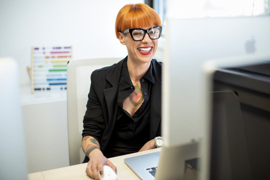 Ragazza tatuata con i capelli rossi e gli occhiali neri con un completo  nero ride nella sua postazione di lavoro