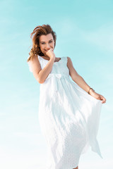 Fototapeta na wymiar low angle view of smiling beautiful girl in white dress against blue sky