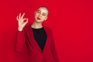 Nice sign showing. Portrait of young caucasian bald woman isolated on red studio background. Beautiful female model in jacket. Human emotions, facial expression, sales, ad concept. Freaky culture.