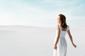 Fototapeta na wymiar back view of smiling beautiful girl in white dress on sandy beach with blue sky