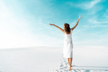 Fototapeta na wymiar back view of beautiful girl in white dress with hands in air on sandy beach with blue sky