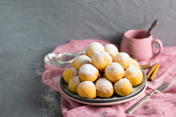 Small homemade curd donuts with icing sugar on a gray concrete background with a pink napkin and a glass of tea