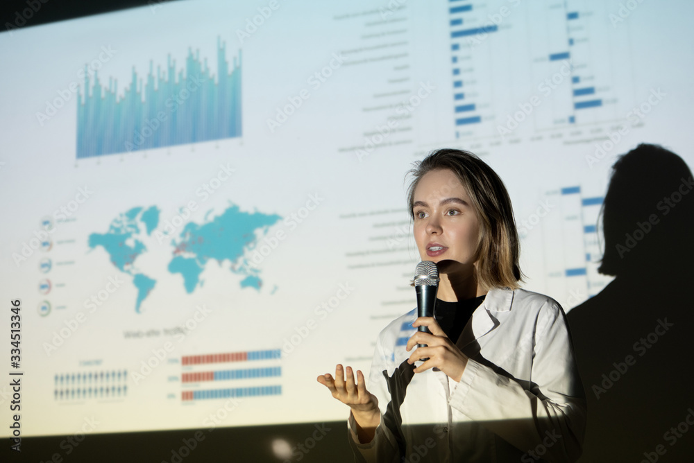 Poster young woman in lab coat gesturing hand and speaking into microphone while persuading investors in pr