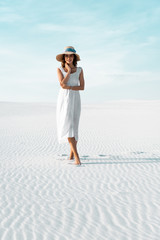 beautiful girl in white dress and straw hat walking on sandy beach with blue sky