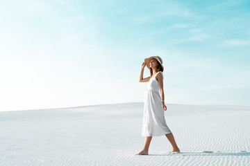 side view of smiling beautiful girl in white dress and straw hat walking on sandy beach with blue...
