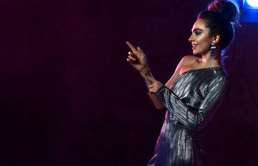 Pretty smiling woman in silver concert dress singer actress is on stage in neon lights pointing fingers at copy space