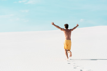 Fototapeta premium back view of man in swim shorts with muscular torso running on sandy beach