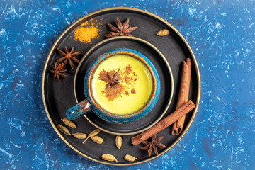 Top view of plates with cup of traditional Indian ayurvedic golden turmeric milk and ingredients on blue background.