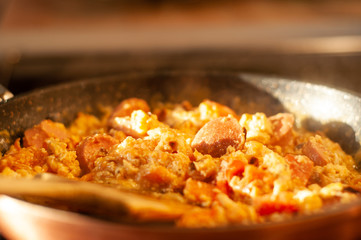 Cooking morning scrambled eggs with sausages, onions and tomatoes in a pan.