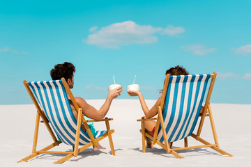 back view of young couple sitting in deck chairs with coconut drinks on sandy beach