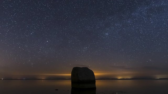 Time lapse of the night sky with the ocean and a huge boulder in the forground