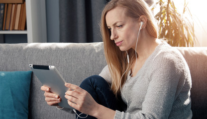 Half-length portrait of blond adult woman using electronic tablet sitting on sofa, side view