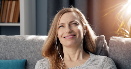 Headshot of smiling blond adult woman relaxing on sofa listening to music via earphones