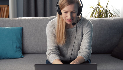 Adult woman in headset with mic sitting on couch at home looking at laptop screen talking to someone