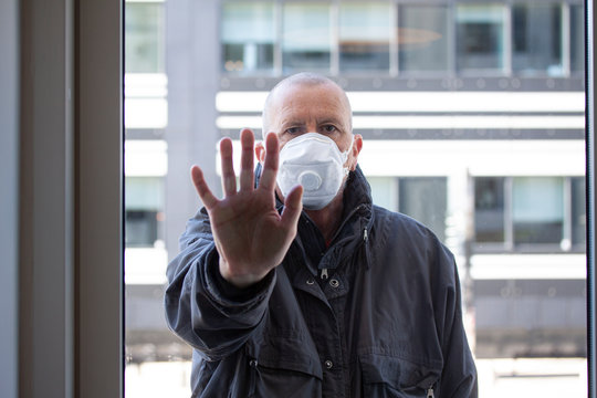 Man With Medical Face Mask Standing Outside A Window Section With His Right Hand Pressed Against Window Glass And Looking Inside. Corona Covid-19 Virus Quarantine Concept.