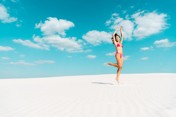 happy beautiful sexy girl in swimsuit jumping with hands in air on sandy beach with blue sky and clouds